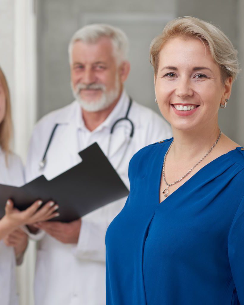 Happy client of private hospital looking at camera and smiling. Doctors wearing in medical uniform holding folder. Old male doctor with stethoscope on neck and young female assistant posing together.